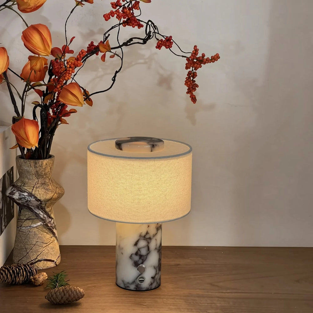 The Marble Drum lamp lit beside autumn flowers, showing the button and USB-C port on the marble base