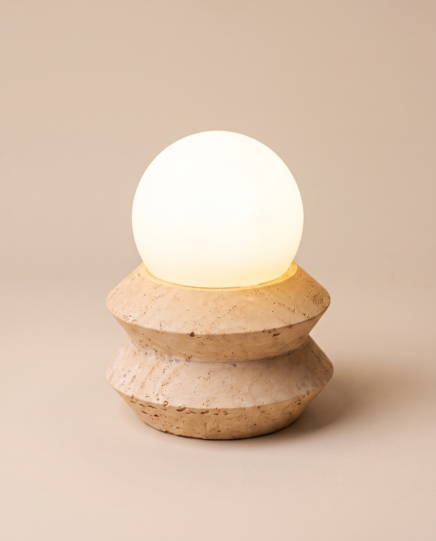 Solace travertine table lamp lit, angled studio view on a warm sand-beige seamless background