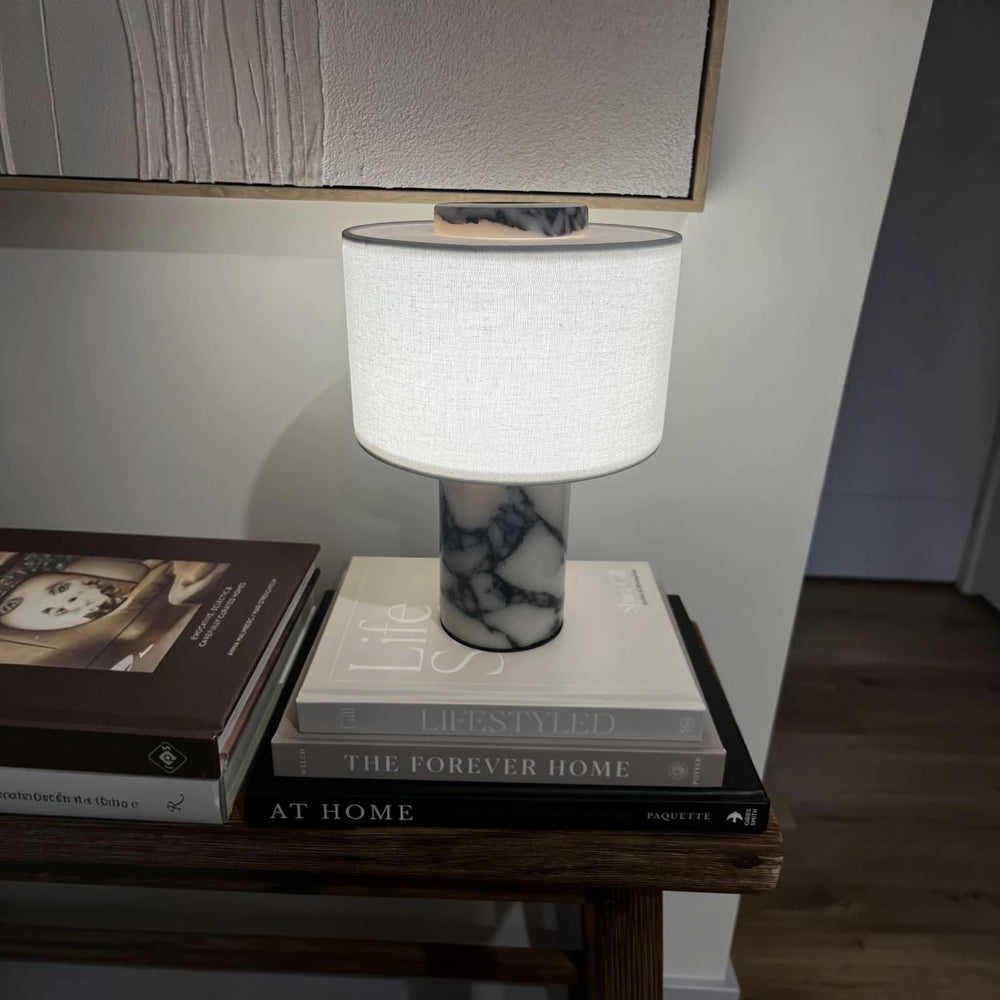 The cordless Marble Drum lamp glowing on a stack of books beneath a framed artwork