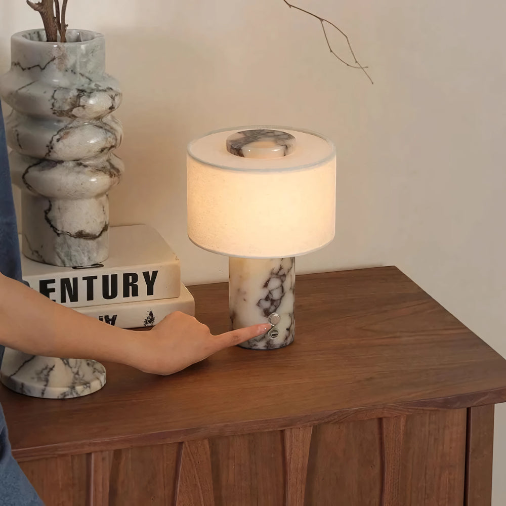 A hand pressing the button on the marble base of the Vensalo Marble Drum lamp, lit on a walnut cabinet
