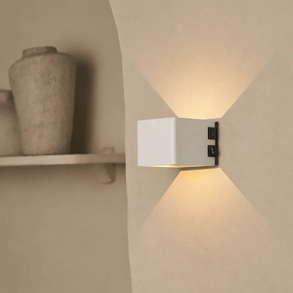 Warm white light from the cordless Cubo sconce washing up and down an alcove wall beside a stone vase