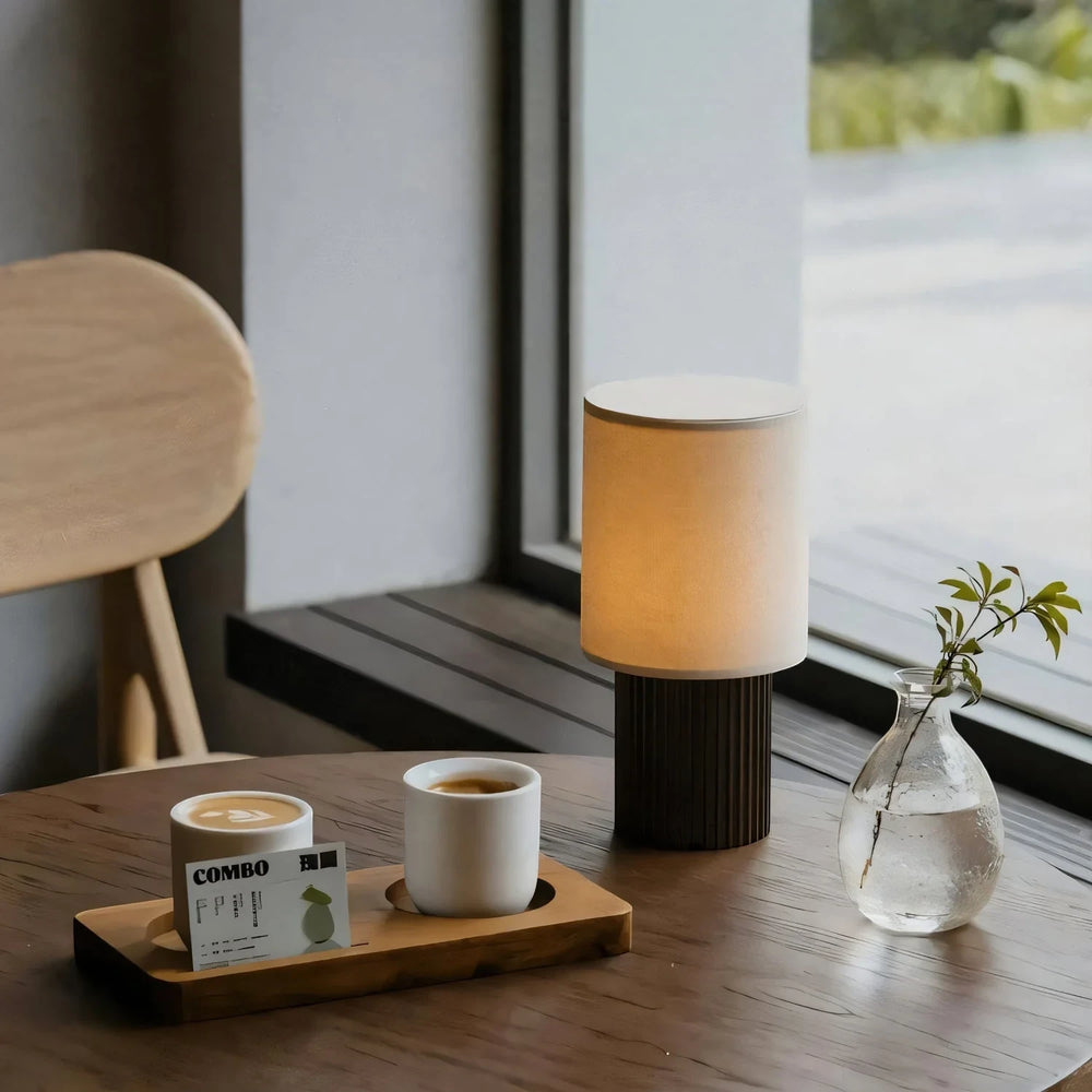 The cordless Solene lamp lit on a wooden table by a window beside two coffee cups